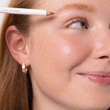 Model positioning an Ere Perez Almond Brow Pencil above her eyebrow to define the brow shape.