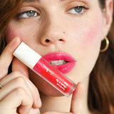A fair-skinned woman with brown hair holds Ere Perez Beetroot Cheek & Lip Tint 4.5ml near her lips, wearing bright pink lipstick, gold hoop earrings, and a gold ring against a soft-focus background.