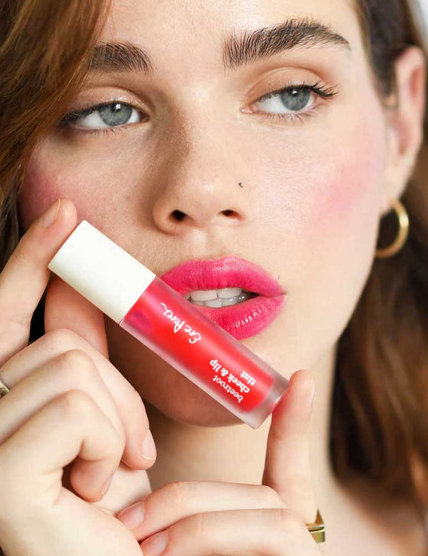 A fair-skinned woman with brown hair holds Ere Perez Beetroot Cheek & Lip Tint 4.5ml near her lips, wearing bright pink lipstick, gold hoop earrings, and a gold ring against a soft-focus background.