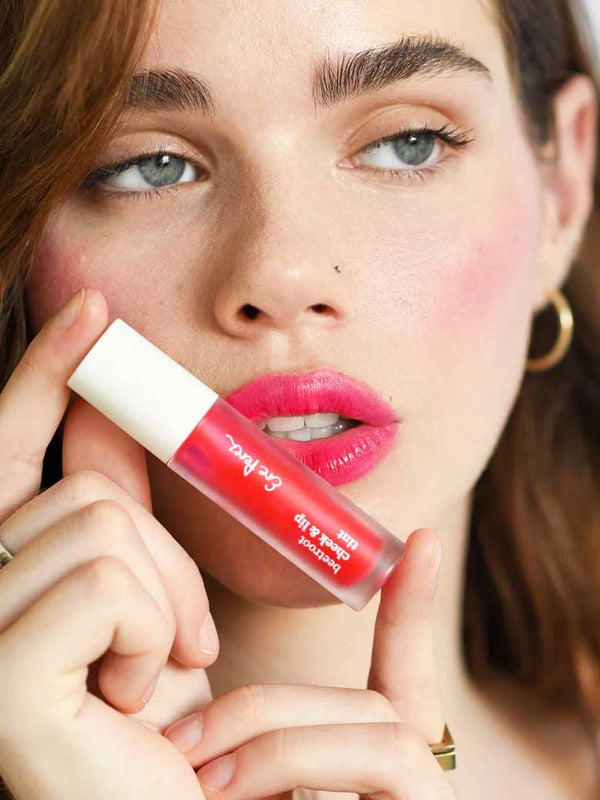 A fair-skinned woman with brown hair holds Ere Perez Beetroot Cheek & Lip Tint 4.5ml near her lips, wearing bright pink lipstick, gold hoop earrings, and a gold ring against a soft-focus background.