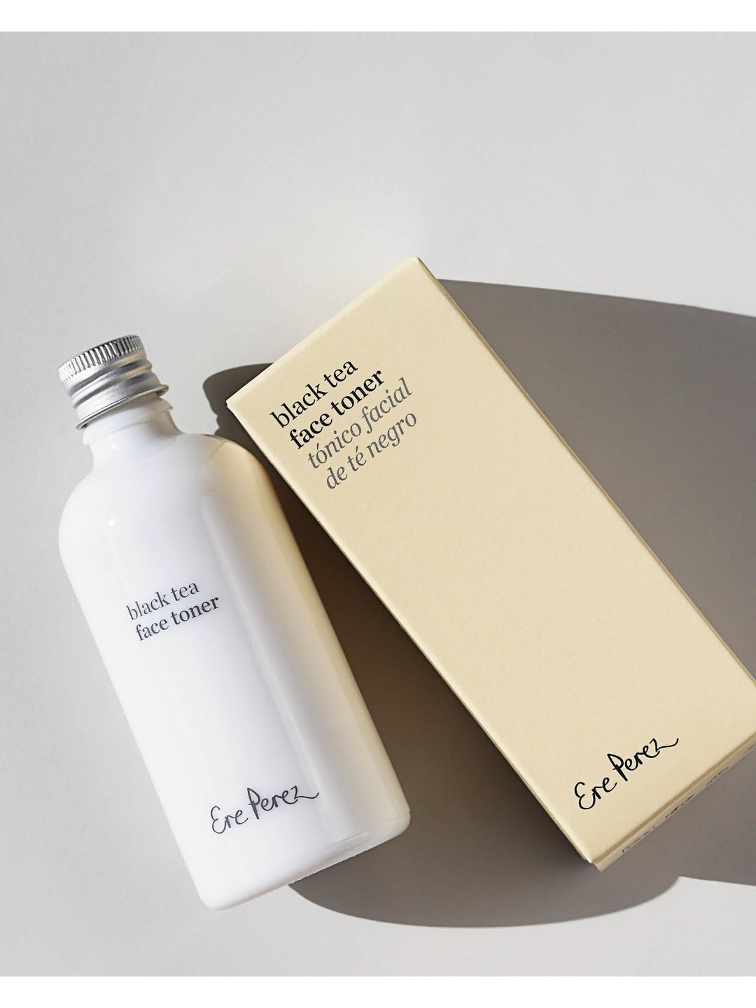 Ere Perez Black Tea Face Toner white bottle and cream carton arranged diagonally on a pale grey surface, with silver cap, printed labels and strong shadows visible.