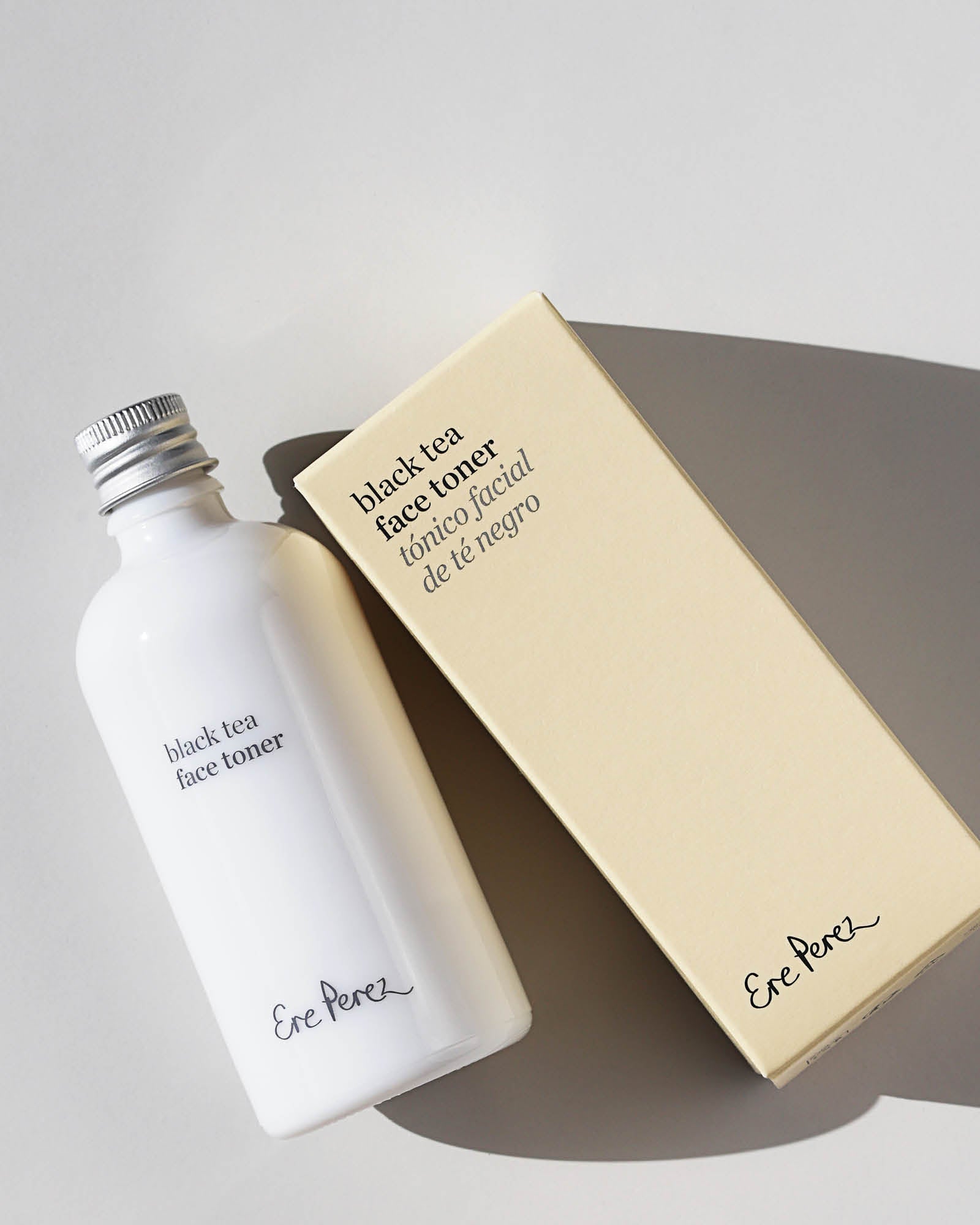 Ere Perez Black Tea Face Toner white bottle and cream carton arranged diagonally on a pale grey surface, with silver cap, printed labels and strong shadows visible.