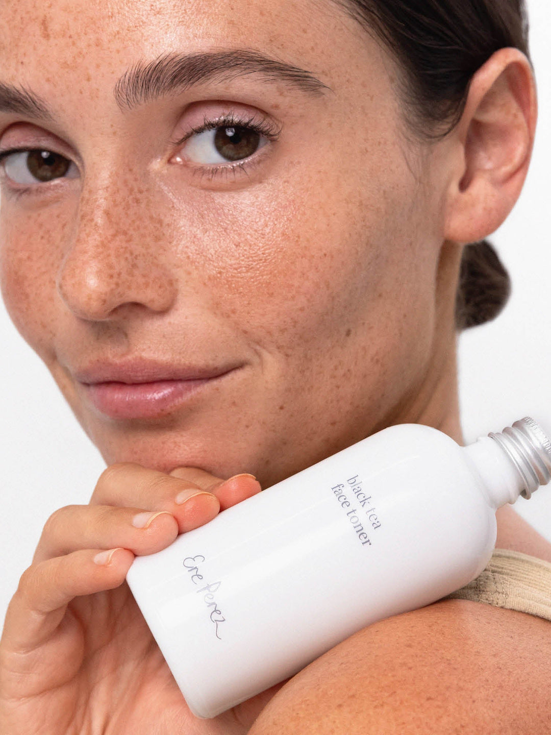 Close-up of a freckled model holding the white Ere Perez Black Tea Face Toner bottle beside her face and shoulder, with its silver cap and grey label visible.