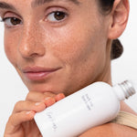 Close-up of a freckled model holding the white Ere Perez Black Tea Face Toner bottle beside her face and shoulder, with its silver cap and grey label visible.