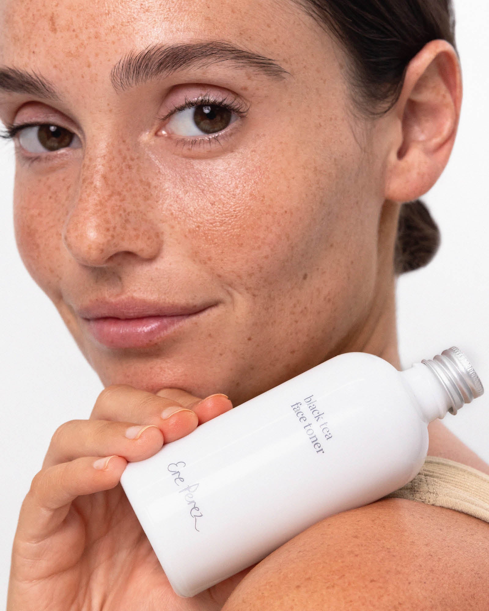 Close-up of a freckled model holding the white Ere Perez Black Tea Face Toner bottle beside her face and shoulder, with its silver cap and grey label visible.