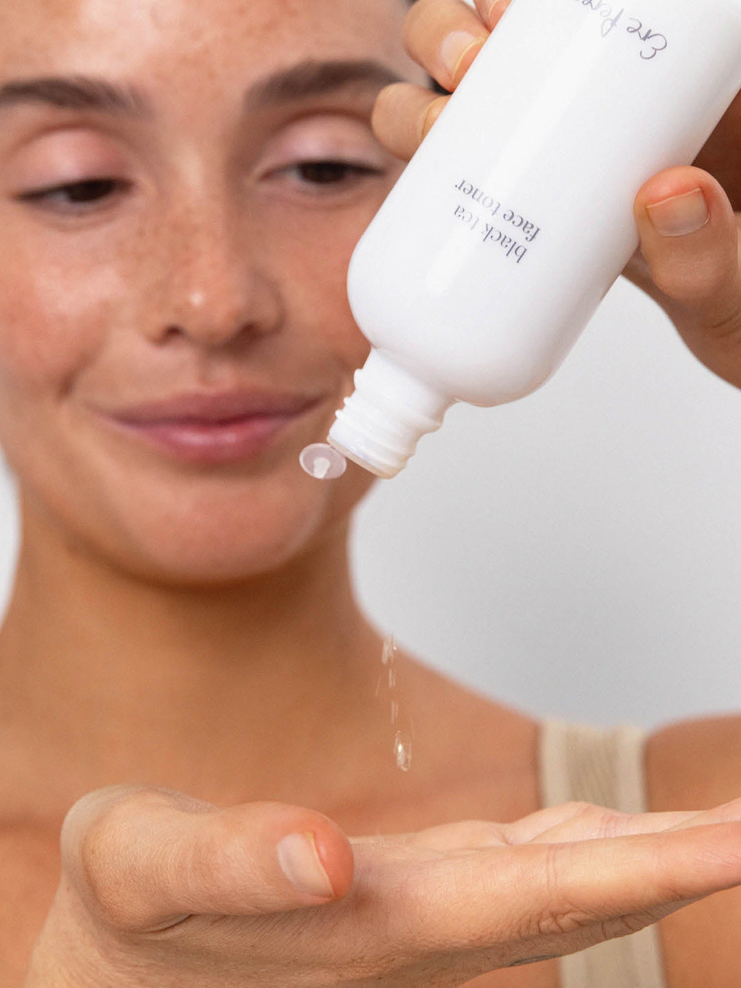 Model tipping an open white Ere Perez Black Tea Face Toner bottle to dispense clear drops of toner into her open palm.