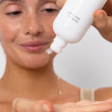 Model tipping an open white Ere Perez Black Tea Face Toner bottle to dispense clear drops of toner into her open palm.