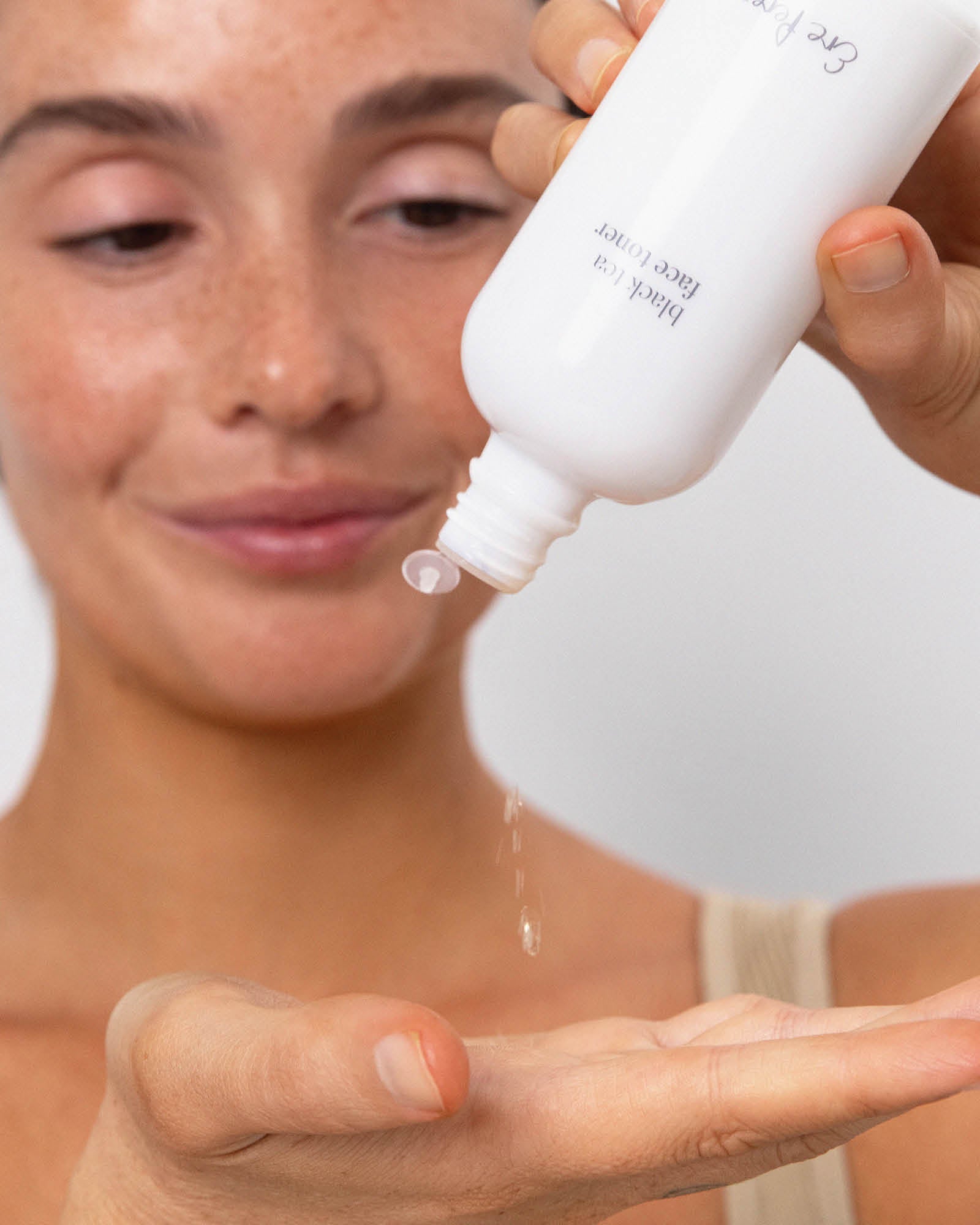 Model tipping an open white Ere Perez Black Tea Face Toner bottle to dispense clear drops of toner into her open palm.