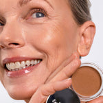 Smiling model wearing Ere Perez Cacao Bronzing Pot in Playa while holding the open bronzing pot beside her face.