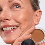 Smiling model wearing Ere Perez Cacao Bronzing Pot in Playa while holding the open bronzing pot beside her face.