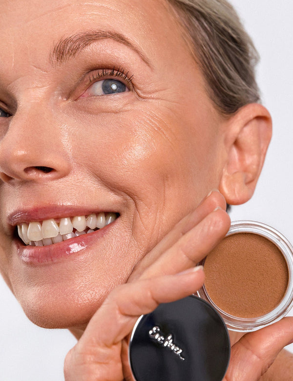 Smiling model wearing Ere Perez Cacao Bronzing Pot in Playa while holding the open bronzing pot beside her face.