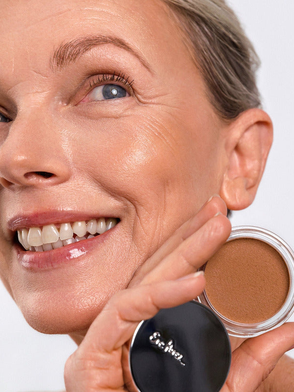 Smiling model wearing Ere Perez Cacao Bronzing Pot in Playa while holding the open bronzing pot beside her face.