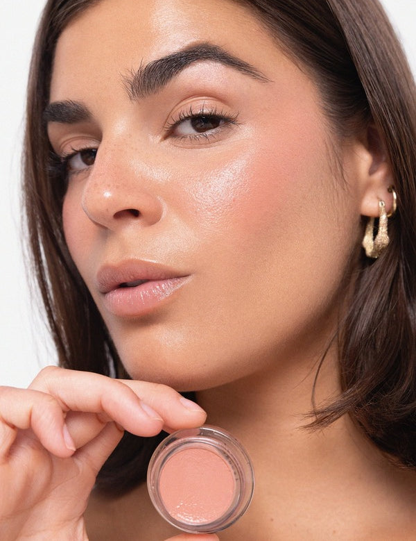 Woman displaying a peach Ere Perez Carrot Colour Pot beside her softly blushed cheek and luminous complexion.