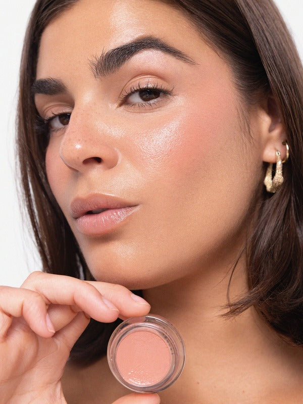 Woman displaying a peach Ere Perez Carrot Colour Pot beside her softly blushed cheek and luminous complexion.