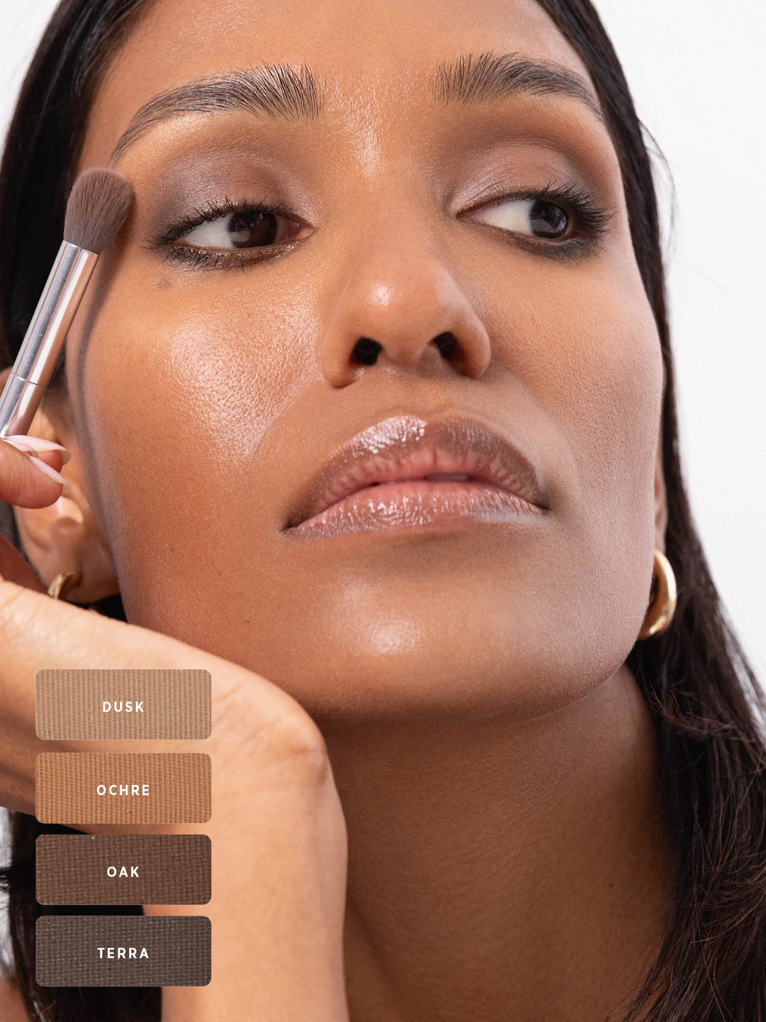 Model blending a smoky brown eye look with a fluffy brush, using the Ere Perez Element Matte palette. Swatches at the lower left identify Dusk, Ochre, Oak and Terra.