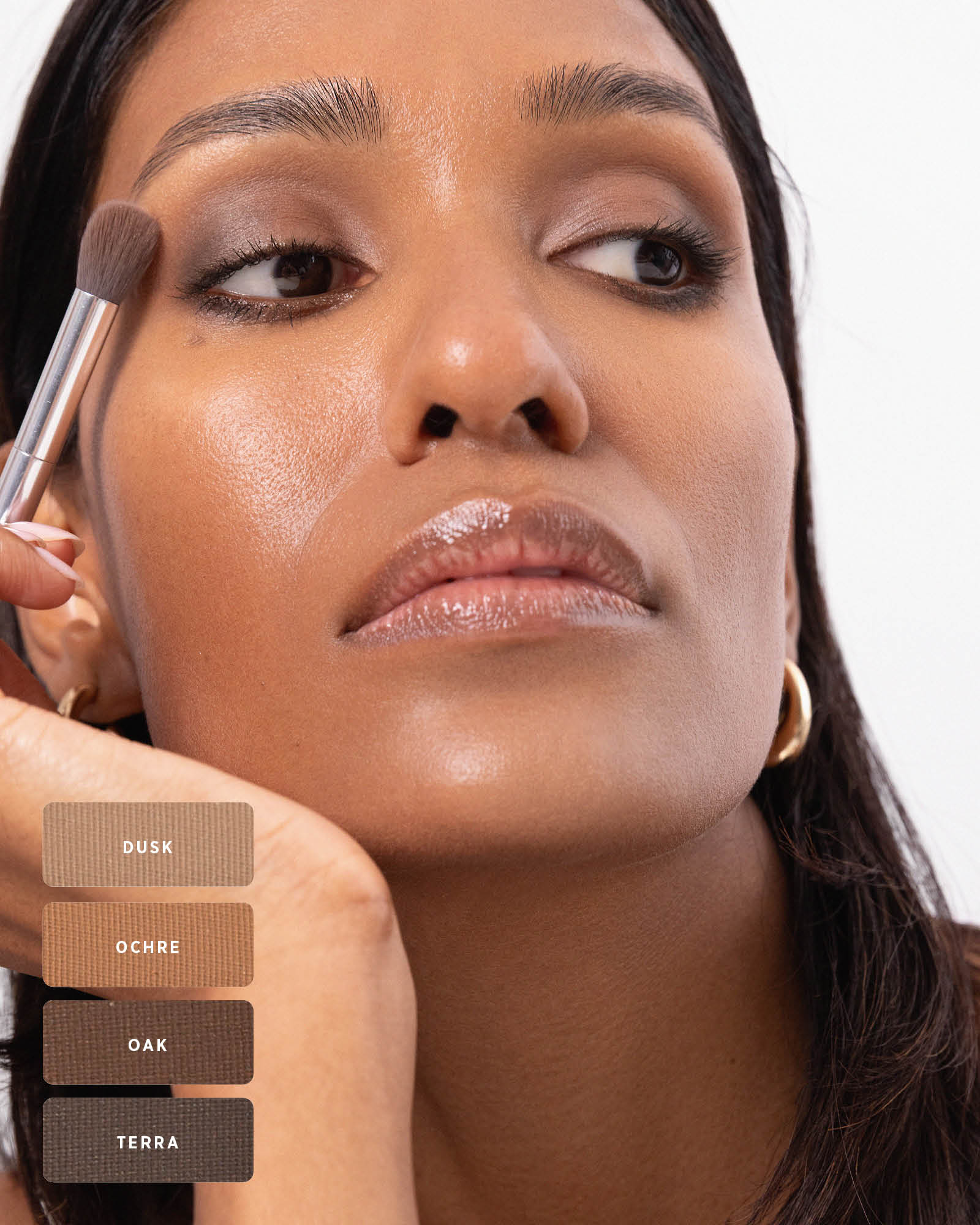 Model blending a smoky brown eye look with a fluffy brush, using the Ere Perez Element Matte palette. Swatches at the lower left identify Dusk, Ochre, Oak and Terra.