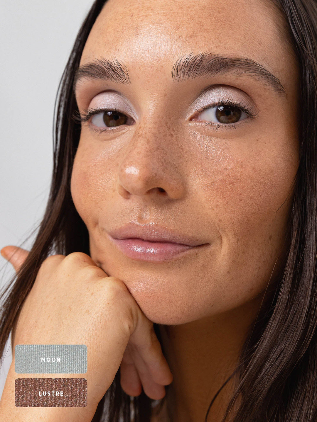 Freckled model resting her chin on her hand, wearing cool silvery shimmer across the eyelids. The Ere Perez Light Shimmer palette shades Moon and Lustre are identified by swatches at the lower left.