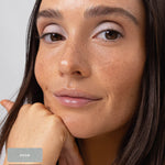 Freckled model resting her chin on her hand, wearing cool silvery shimmer across the eyelids. The Ere Perez Light Shimmer palette shades Moon and Lustre are identified by swatches at the lower left.