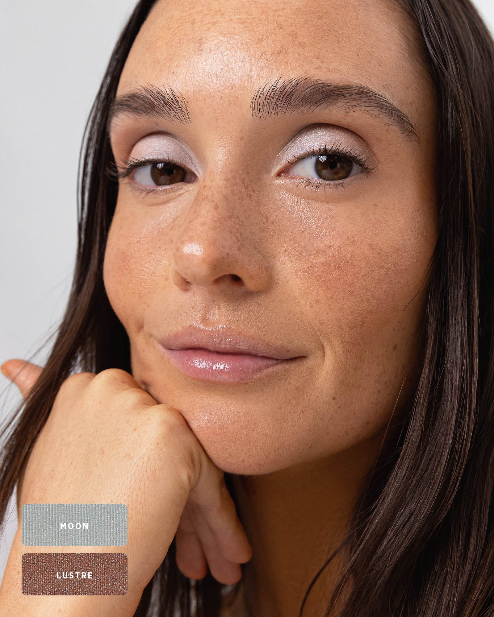 Freckled model resting her chin on her hand, wearing cool silvery shimmer across the eyelids. The Ere Perez Light Shimmer palette shades Moon and Lustre are identified by swatches at the lower left.