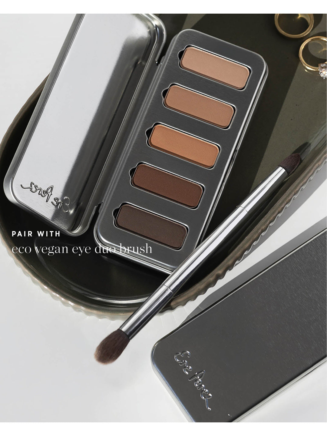 Ere Perez Eco Vegan Eye Duo Brush resting diagonally across a metal tray beside an open Element Matte Chamomile Eye Palette, with both brush heads visible. Text reads 'Pair with eco vegan eye duo brush'.