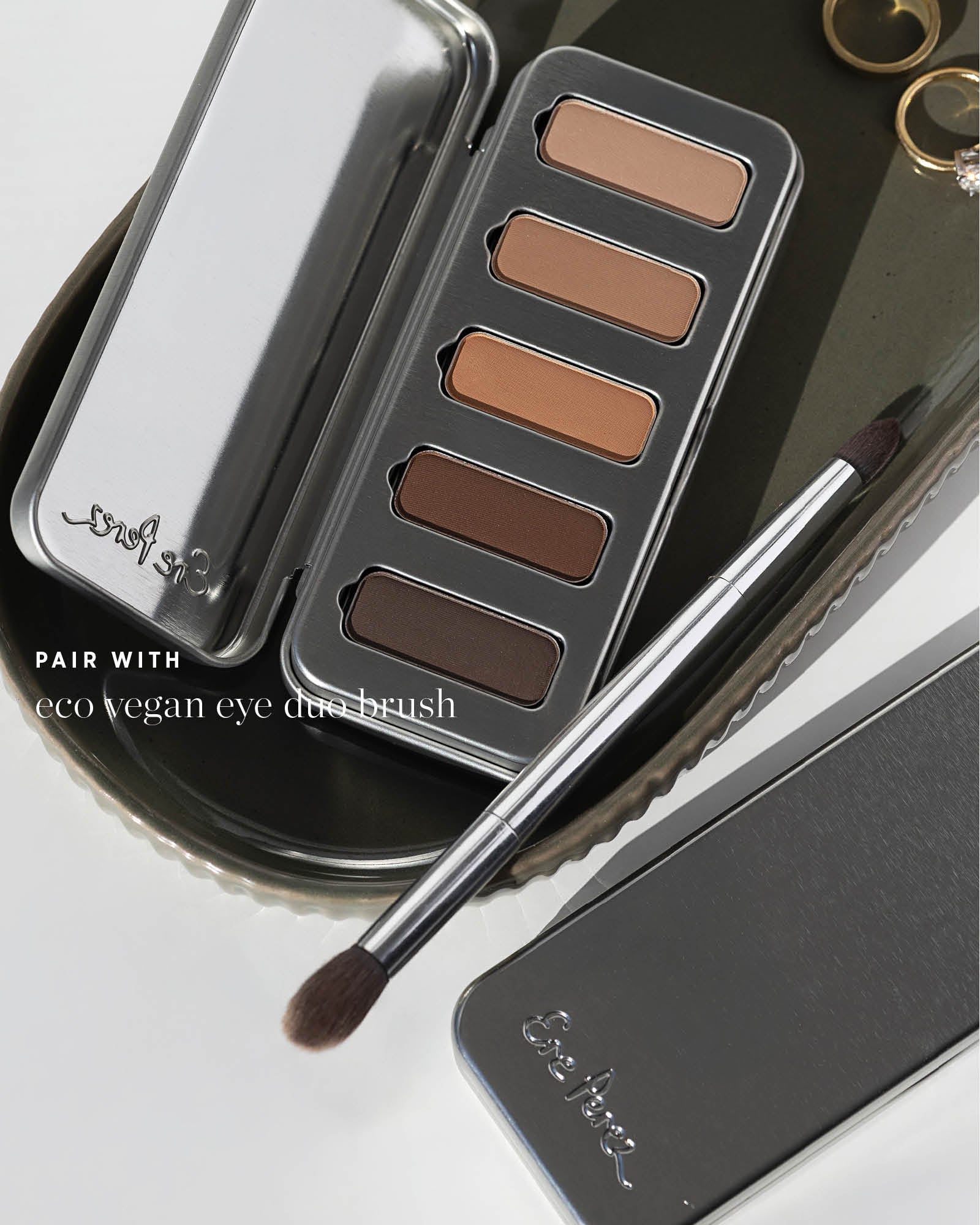 Ere Perez Eco Vegan Eye Duo Brush resting diagonally across a metal tray beside an open Element Matte Chamomile Eye Palette, with both brush heads visible. Text reads 'Pair with eco vegan eye duo brush'.