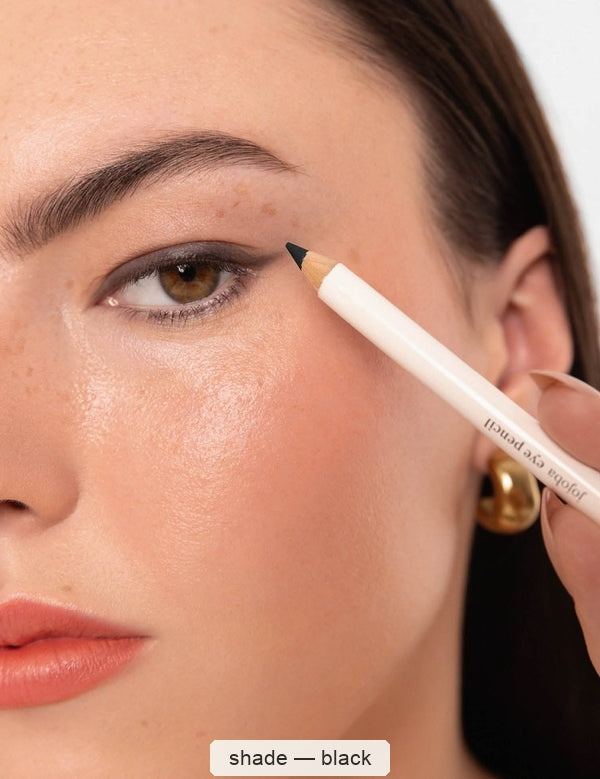 Close-up of a woman applying a white Ere Perez Jojoba Eye Pencil to the outer corner of her eye, creating a dark wing along the upper lash line; the caption names the shade Black.