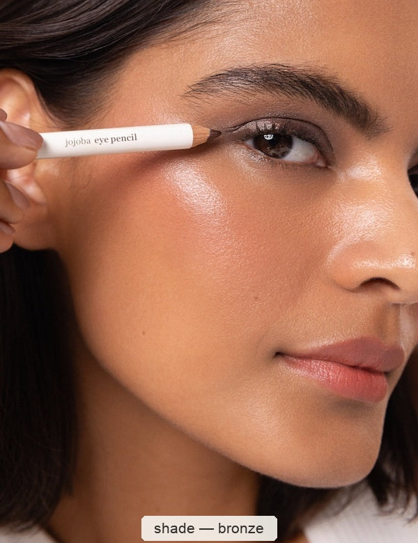 A woman applies a white Ere Perez Jojoba Eye Pencil along her upper lash line, creating a soft bronze-brown outline; the caption names the shade Bronze.
