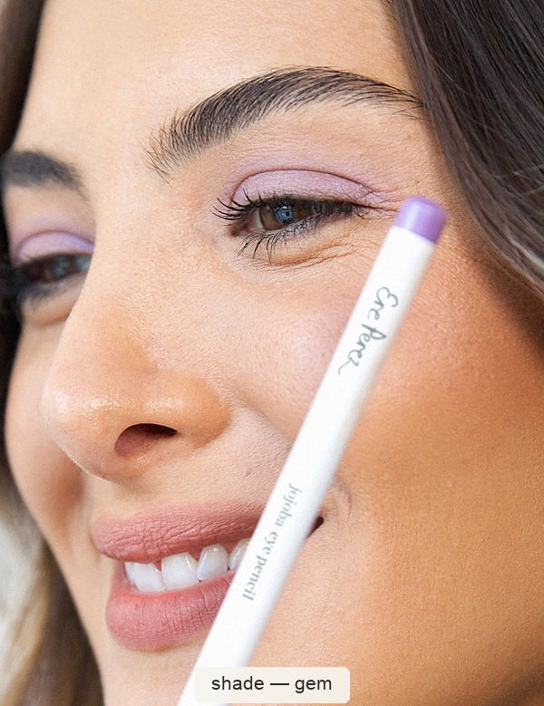 Close-up of a smiling woman with lavender eyeliner across her upper eyelid, holding a white Ere Perez Jojoba Eye Pencil with a purple end cap beside her cheek; the caption names the shade Gem.