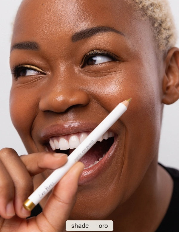 A smiling woman with short light-blonde hair holds a white Ere Perez Jojoba Eye Pencil beside her face, showing gold eyeliner along her eyes; the caption names the shade Oro.