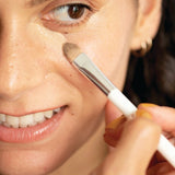 Close-up of a woman applying Ere Perez Lychee Crème Corrector beneath her eye with a small flat makeup brush.