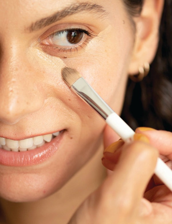 Close-up of a woman applying Ere Perez Lychee Crème Corrector beneath her eye with a small flat makeup brush.