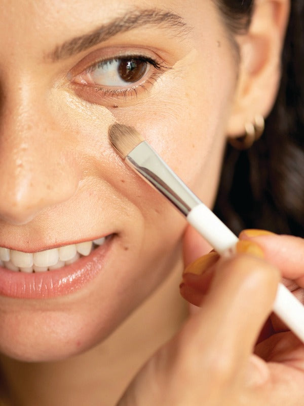 Close-up of a woman applying Ere Perez Lychee Crème Corrector beneath her eye with a small flat makeup brush.