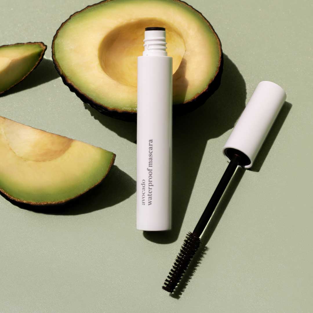 A tube of avocado waterproof mascara with the wand open, placed next to a halved avocado and avocado slices on a light green surface.