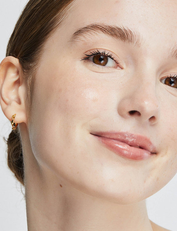 Close-up portrait of a woman wearing Ere Perez Oat Milk Foundation in Nougat, with an even light-beige complexion, faint freckles across her cheeks and softly glossy pink lips.