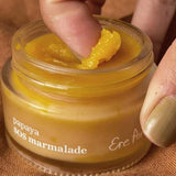 A close-up of fingers scooping bright yellow Ere Perez Papaya SOS Marmalade from a small glass jar against a warm-toned background.