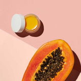 A small open jar of Ere Perez Papaya SOS Marmalade, a yellow-orange multipurpose balm, sits beside a halved papaya with black seeds on a split pink background.