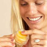 A smiling person with light skin, blonde hair, and a shell ring dips a finger into a small jar of Ere Perez Papaya SOS Marmalade healing balm.