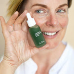 Smiling woman holding an Ere Perez Quandong Green Booster Serum bottle beside her face, showing the green bottle and white dropper cap.