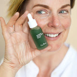 Smiling woman holding an Ere Perez Quandong Green Booster Serum bottle beside her face, showing the green bottle and white dropper cap.