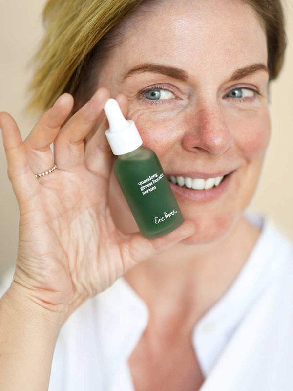 Smiling woman holding an Ere Perez Quandong Green Booster Serum bottle beside her face, showing the green bottle and white dropper cap.
