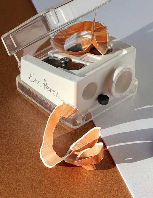 The Ere Perez Eco Duo Sharpener, in white with carbon steel blades, sits open on a brown and white surface in sunlight, filled with peach pencil shavings. Its durable design is ideal for sharpening makeup pencils.