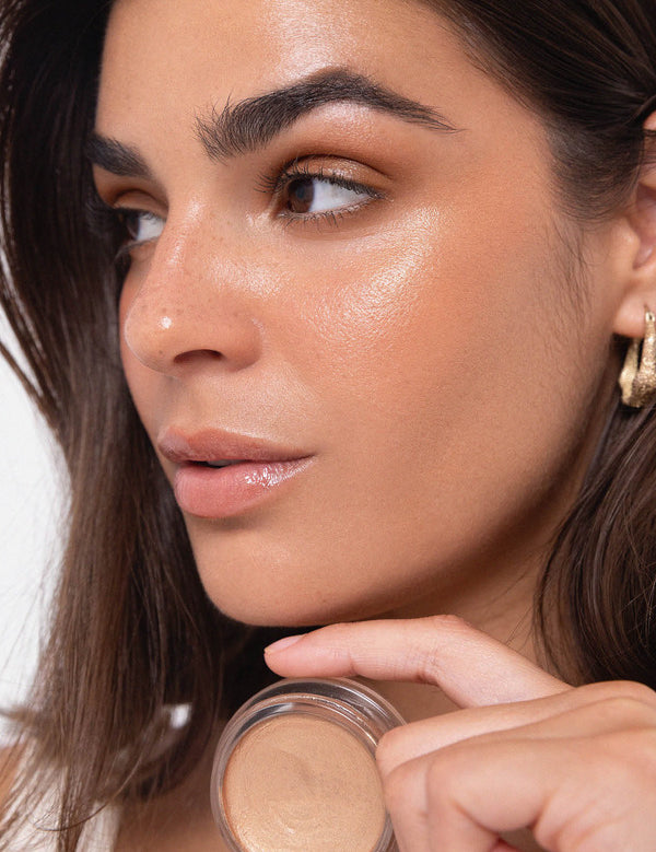 Model wearing luminous Ere Perez Vanilla Highlighter Balm while holding the open balm near her shoulder.