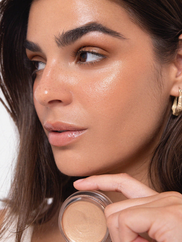 Model wearing luminous Ere Perez Vanilla Highlighter Balm while holding the open balm near her shoulder.