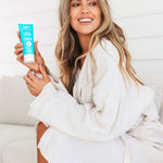 A smiling woman in a white bathrobe sits sideways on a pale seat, holding a turquoise Ethical Zinc sunscreen tube upright beside her long wavy blonde hair.