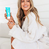 A smiling woman in a white bathrobe sits sideways on a pale seat, holding a turquoise Ethical Zinc sunscreen tube upright beside her long wavy blonde hair.