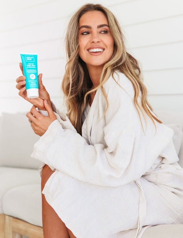 A smiling woman in a white bathrobe sits sideways on a pale seat, holding a turquoise Ethical Zinc sunscreen tube upright beside her long wavy blonde hair.