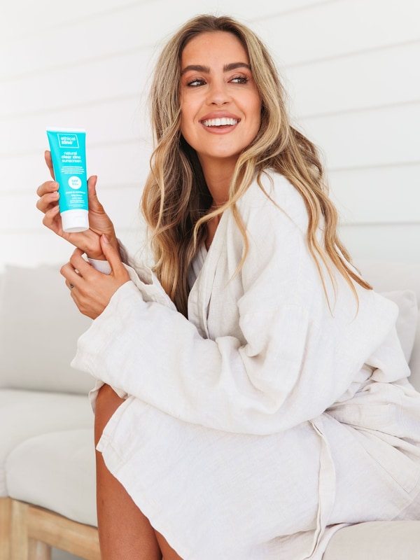 A smiling woman in a white bathrobe sits sideways on a pale seat, holding a turquoise Ethical Zinc sunscreen tube upright beside her long wavy blonde hair.