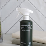Dark green Euclove Home Spray bottle with a white trigger sprayer on a pale counter beside folded towels, with diagonal white tiles and green foliage behind.