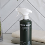 Dark green Euclove Home Spray bottle with a white trigger sprayer on a pale counter beside folded towels, with diagonal white tiles and green foliage behind.