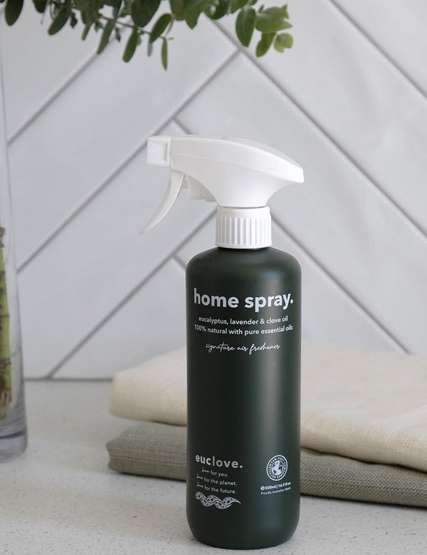 Dark green Euclove Home Spray bottle with a white trigger sprayer on a pale counter beside folded towels, with diagonal white tiles and green foliage behind.